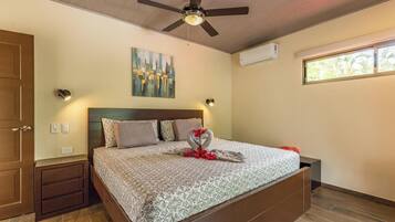 A bedroom with a bed, a ceiling fan, an air conditioning unit, a window, and a painting on the wall.