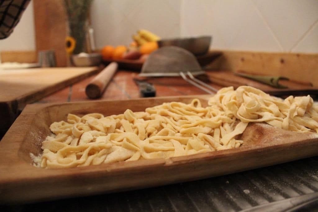 A wooden cutting board with pasta on it, a rolling pin, and kitchen utensils in the background.