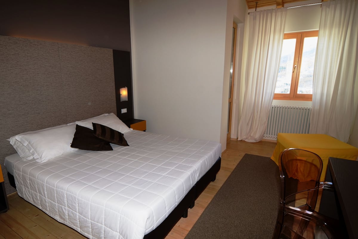 A neatly made bed with white bedding and two pillows, a wooden nightstand with a lamp, a small table with a yellow tablecloth, and a chair.