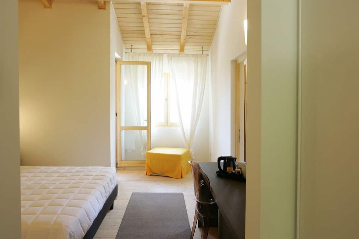 A bedroom with a bed, a small table with a kettle, and a chair.