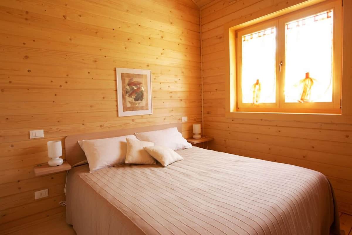 A wooden cabin bedroom with a bed, pillows, a nightstand, a framed picture, and a window with two figures silhouetted against the light.
