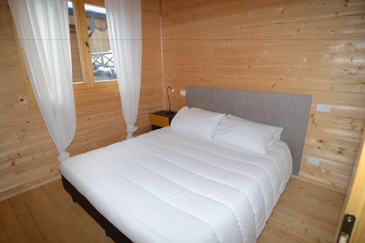 A wooden cabin bedroom with a bed, curtains, a nightstand, and a window.