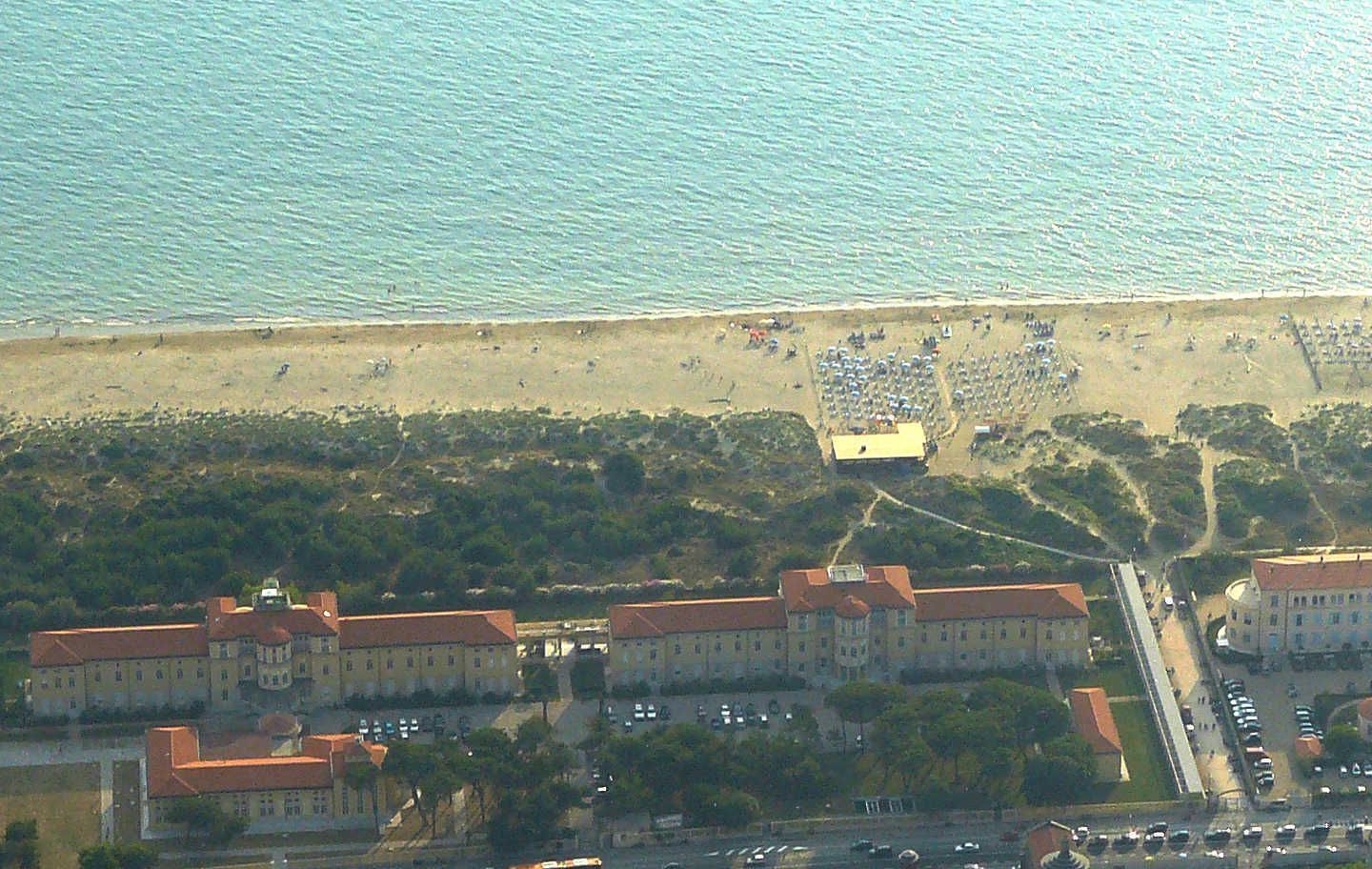An aerial view of a coastal area with a beach, a large building complex, and a parking area.