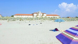 Een strand met een groot gebouw op de achtergrond, een gestreept strandparasol en een strandstoel.