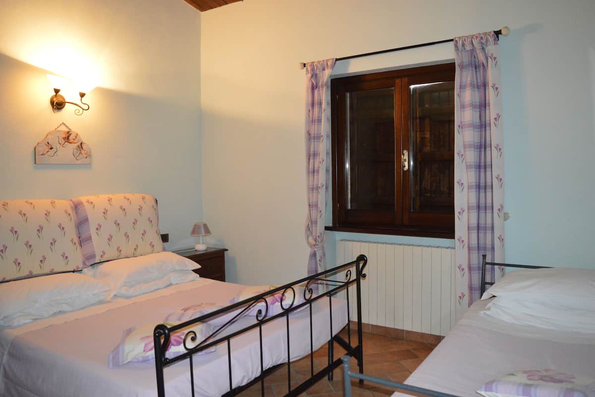 A bedroom with two beds, a window with curtains, a wall-mounted light fixture, and a small bedside table with a lamp.