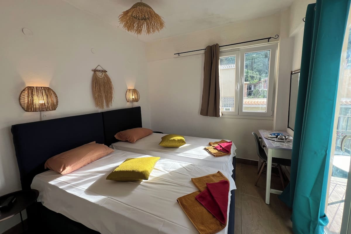 A neatly made bed with white bedding and multiple pillows, a small table with a laptop, and a window with curtains.