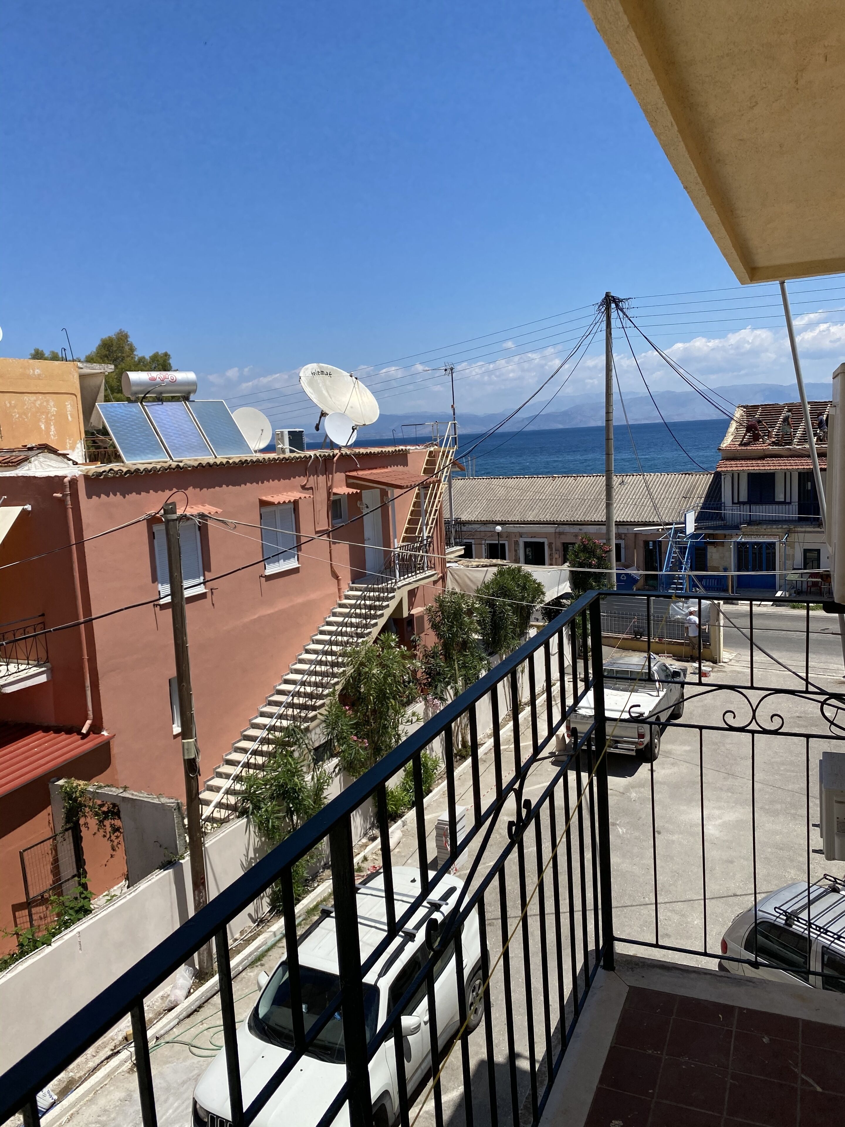 A residential area with a balcony overlooking a street, solar panels on a building, and a clear view of the sea.
