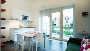 Cocina moderna con armarios blancos y un microondas, comedor con mesa y sillas, y sofá verde con cojín floral.