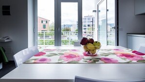 Una mesa de comedor con mantel floral, un cuenco de fruta y vistas a edificios y vegetación a través de amplios ventanales.