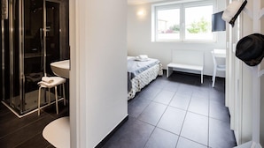 Un baño moderno con cabina de ducha de vidrio, lavamanos y retrete. Hay una cama con cabecero blanco y una ventana que proporciona luz natural.