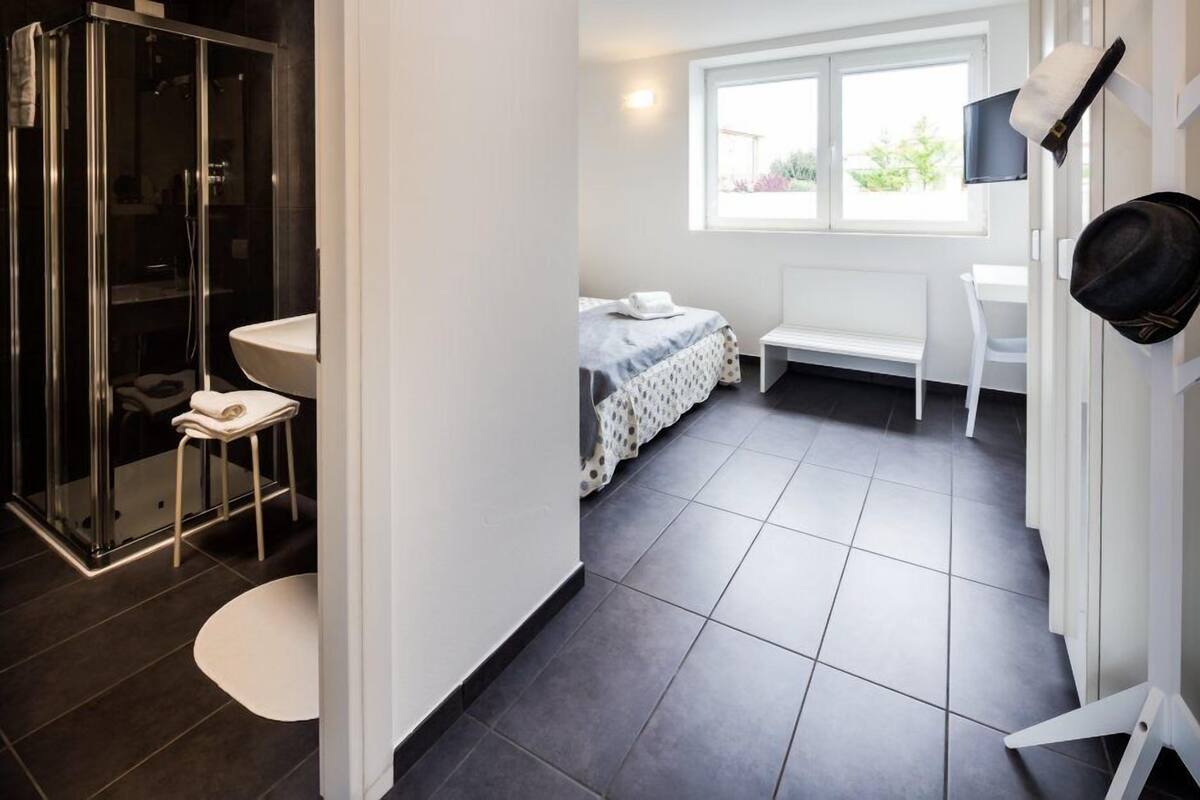 A modern bathroom with a glass shower enclosure, a sink, and a toilet. There is a bed with a white headboard and a window providing natural light.