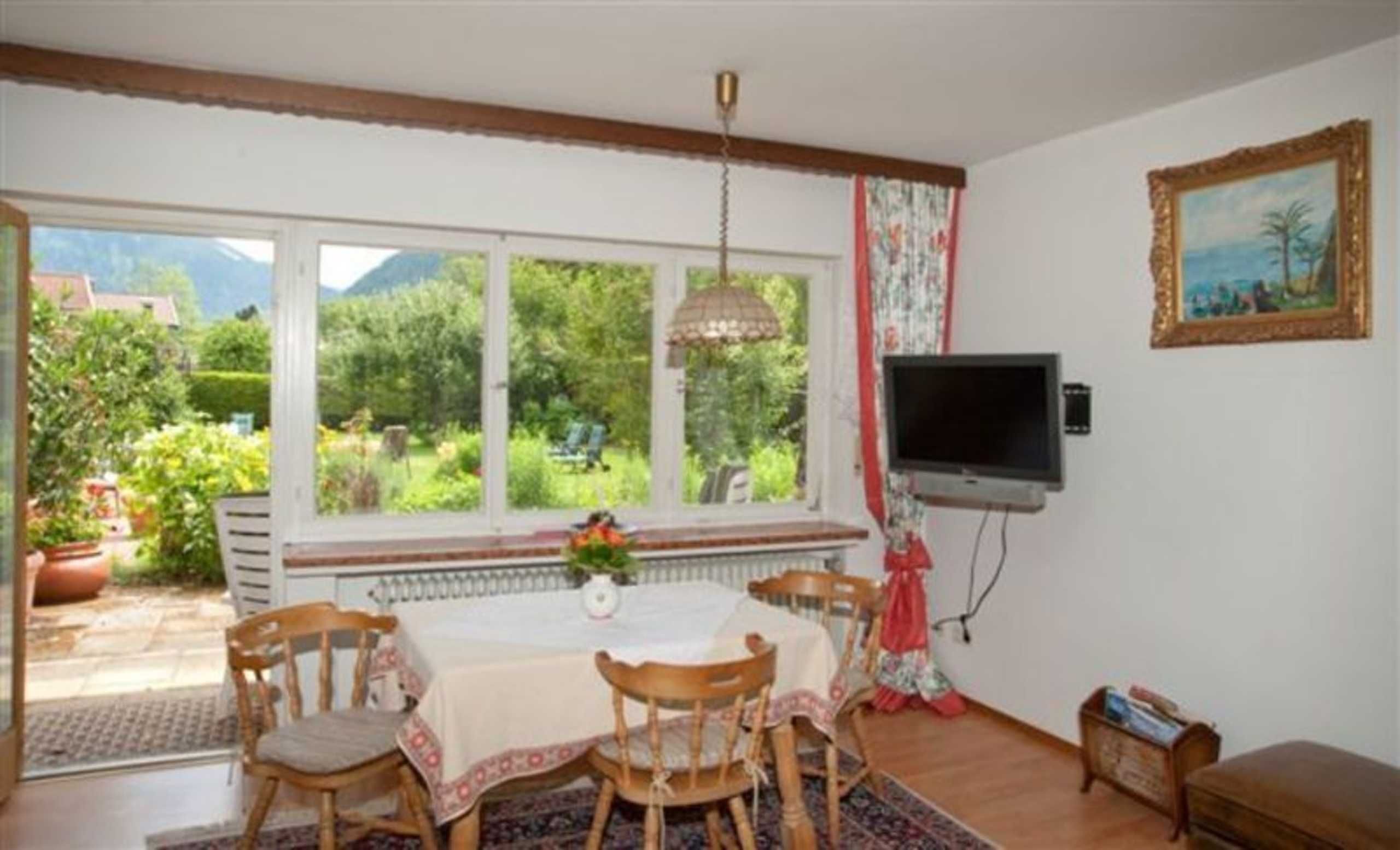 A dining area with a table and chairs, a TV mounted on the wall, and a window with a view of greenery and mountains.