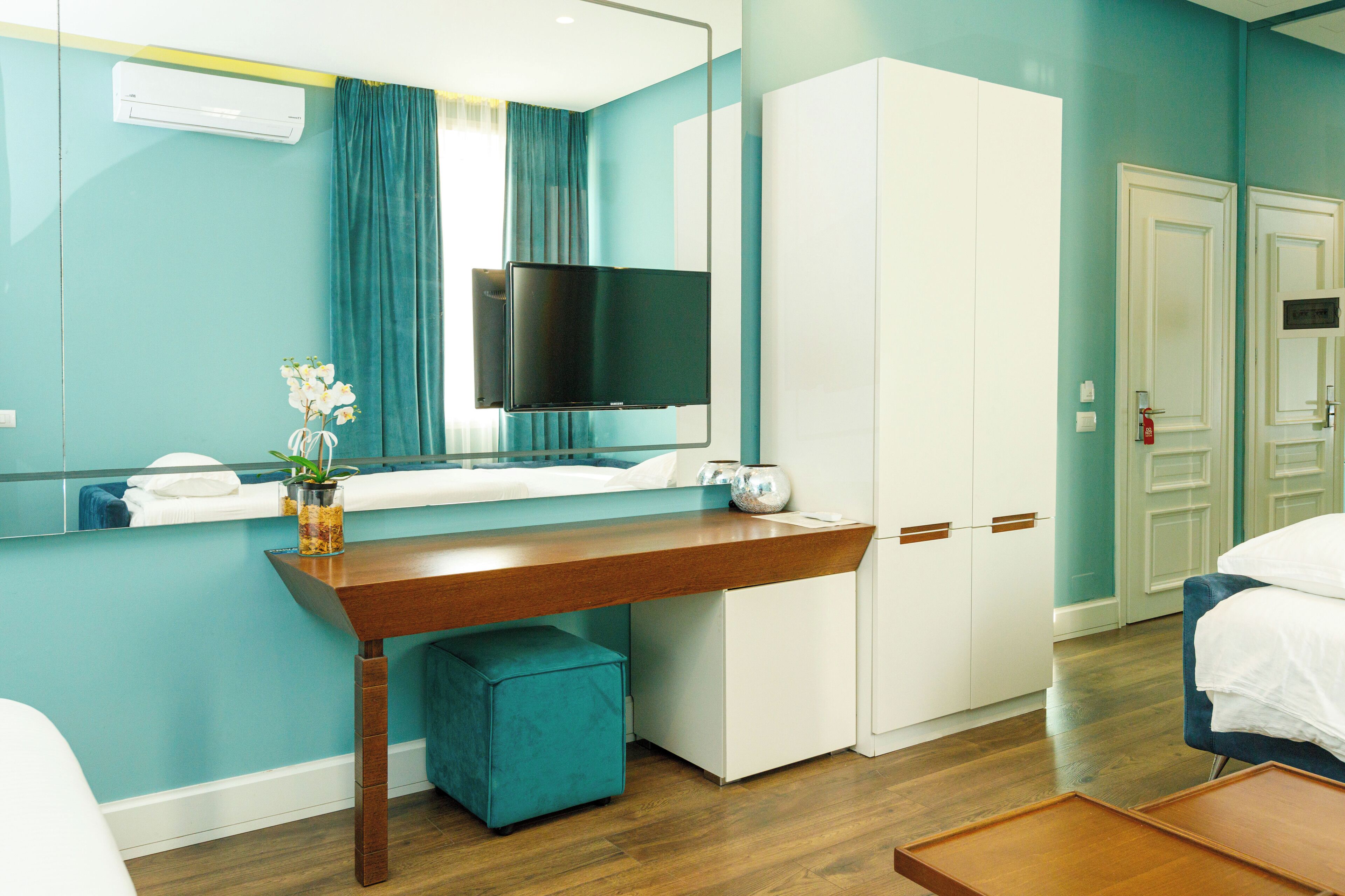 A hotel room with a wooden desk, a flat-screen TV, a bed, a wardrobe, and a mirror.