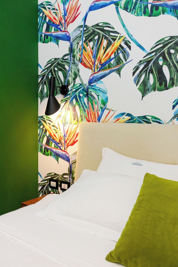 Ein Bett mit weißer Bettwäsche und einem grünen Kopfkissen, eine Wand mit tropischem Blattmuster sowie eine schwarze Nachttischlampe.