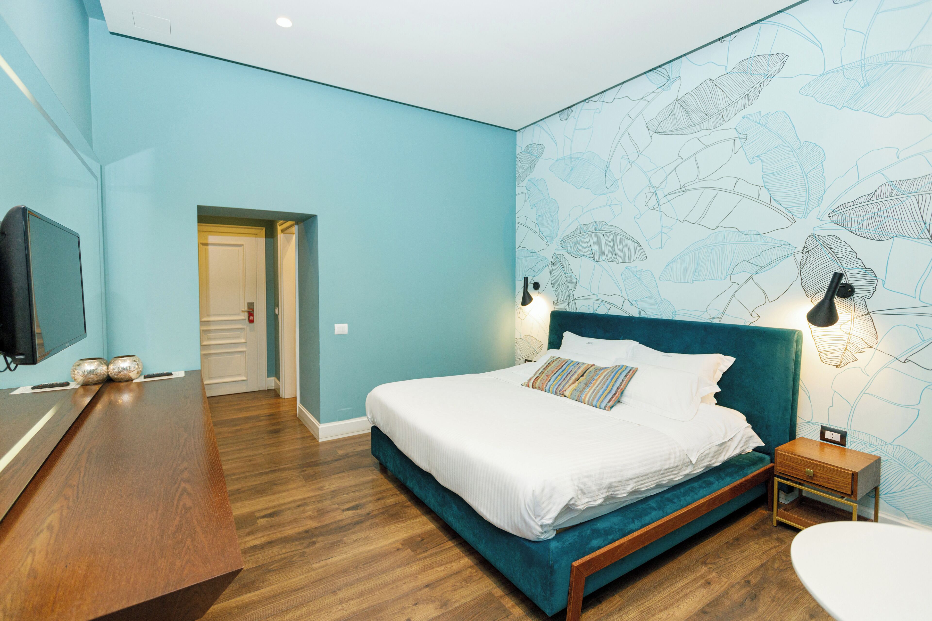 A bedroom with a teal-colored bed, a wooden dresser, and a wall with leaf patterns.