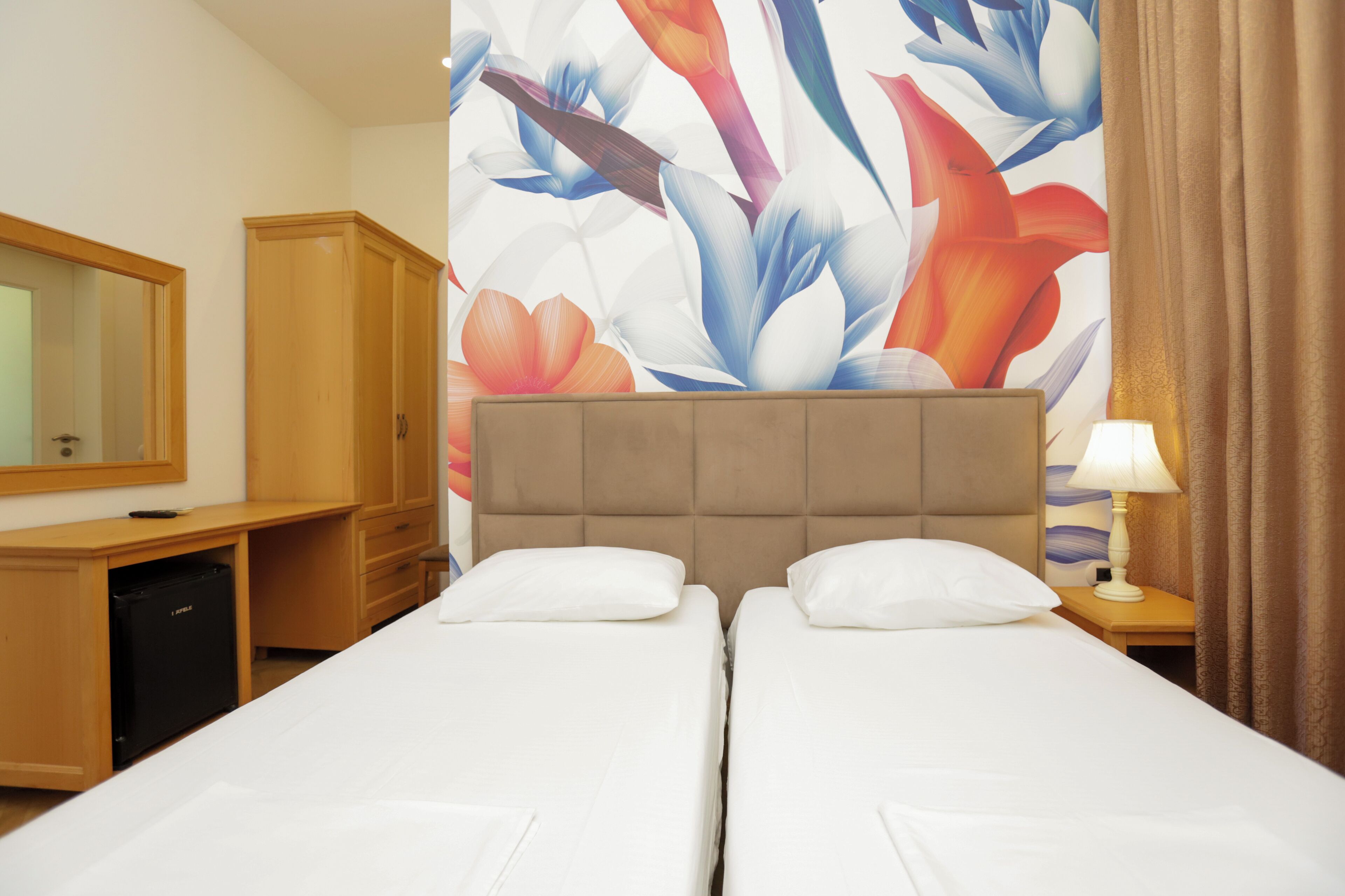Hotellihuone, jossa on kaksi sänkyä, puinen työpöytä, minibaari ja suuri, kukkakuviollinen tapetti.