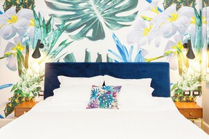 Una cama con edredón blanco, almohada decorativa, cabecera azul oscuro y un mural de flores y plantas tropicales.