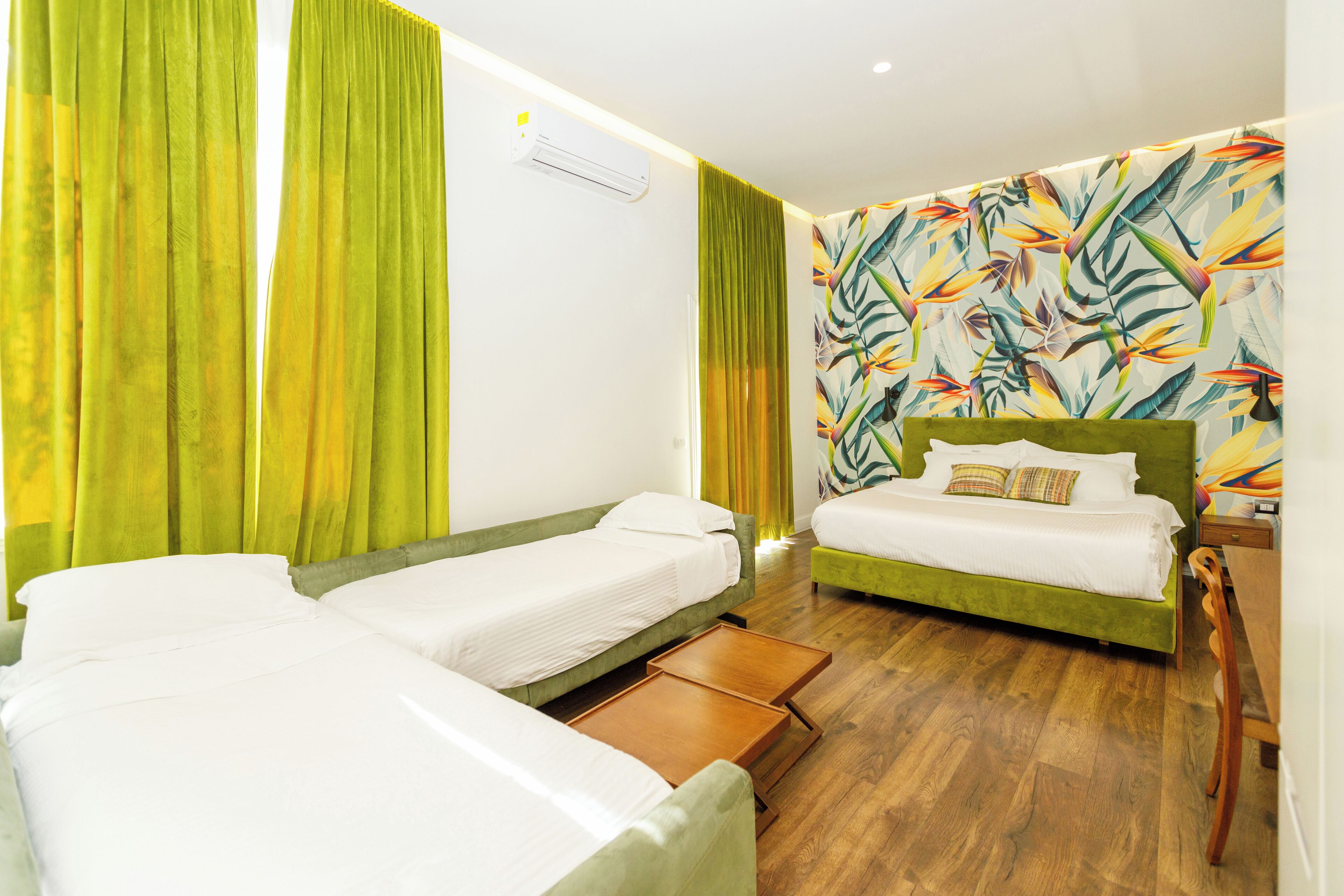 A hotel room with two single beds, a green sofa, a wooden side table, and a colorful tropical wallpaper.