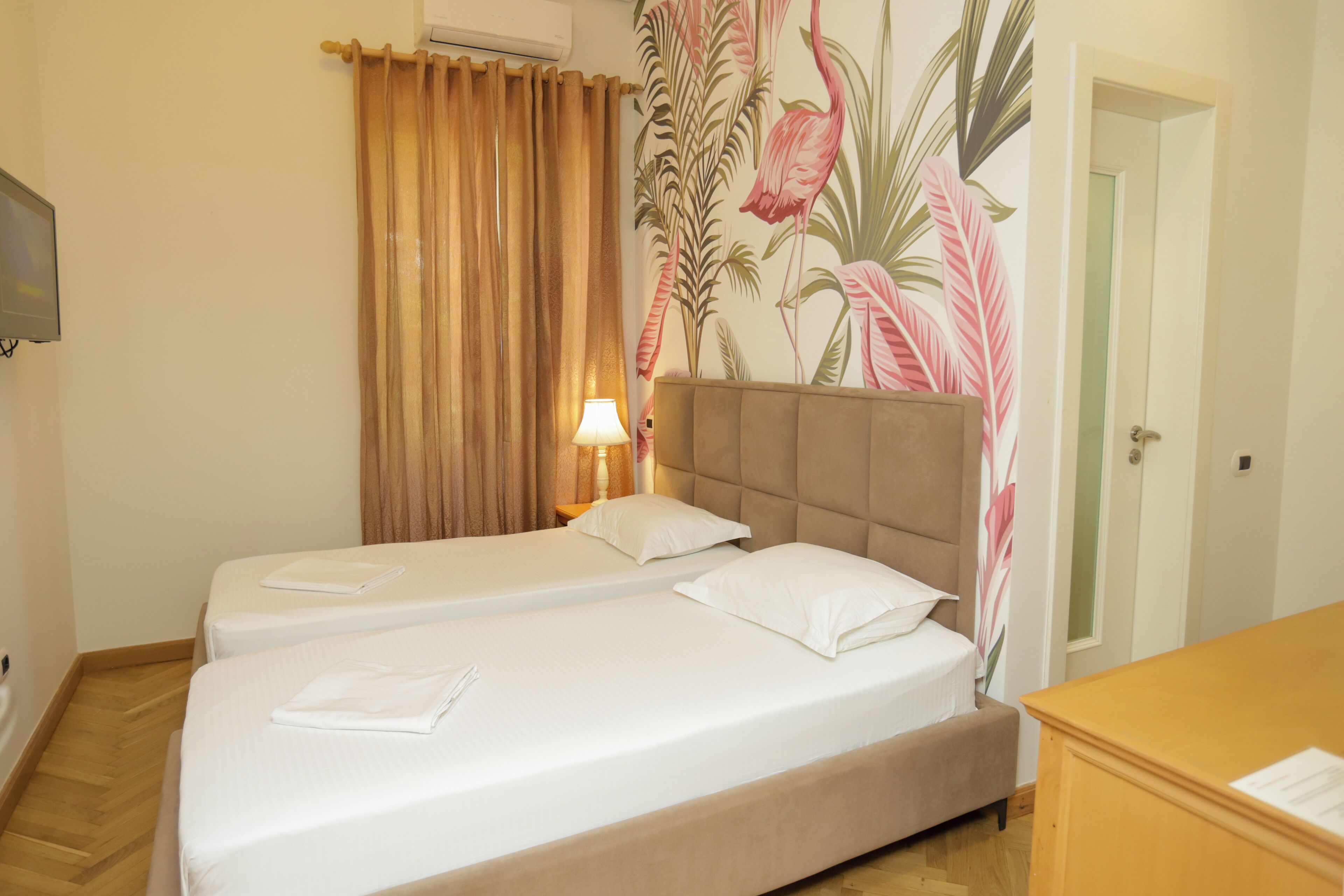 A hotel room with two beds, a wooden desk, a lamp, and a wall with a tropical mural.