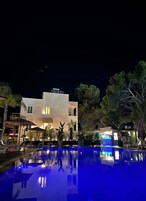 Vista nocturna de un hotel con piscina, mobiliario exterior y un edificio con ventanas iluminadas.