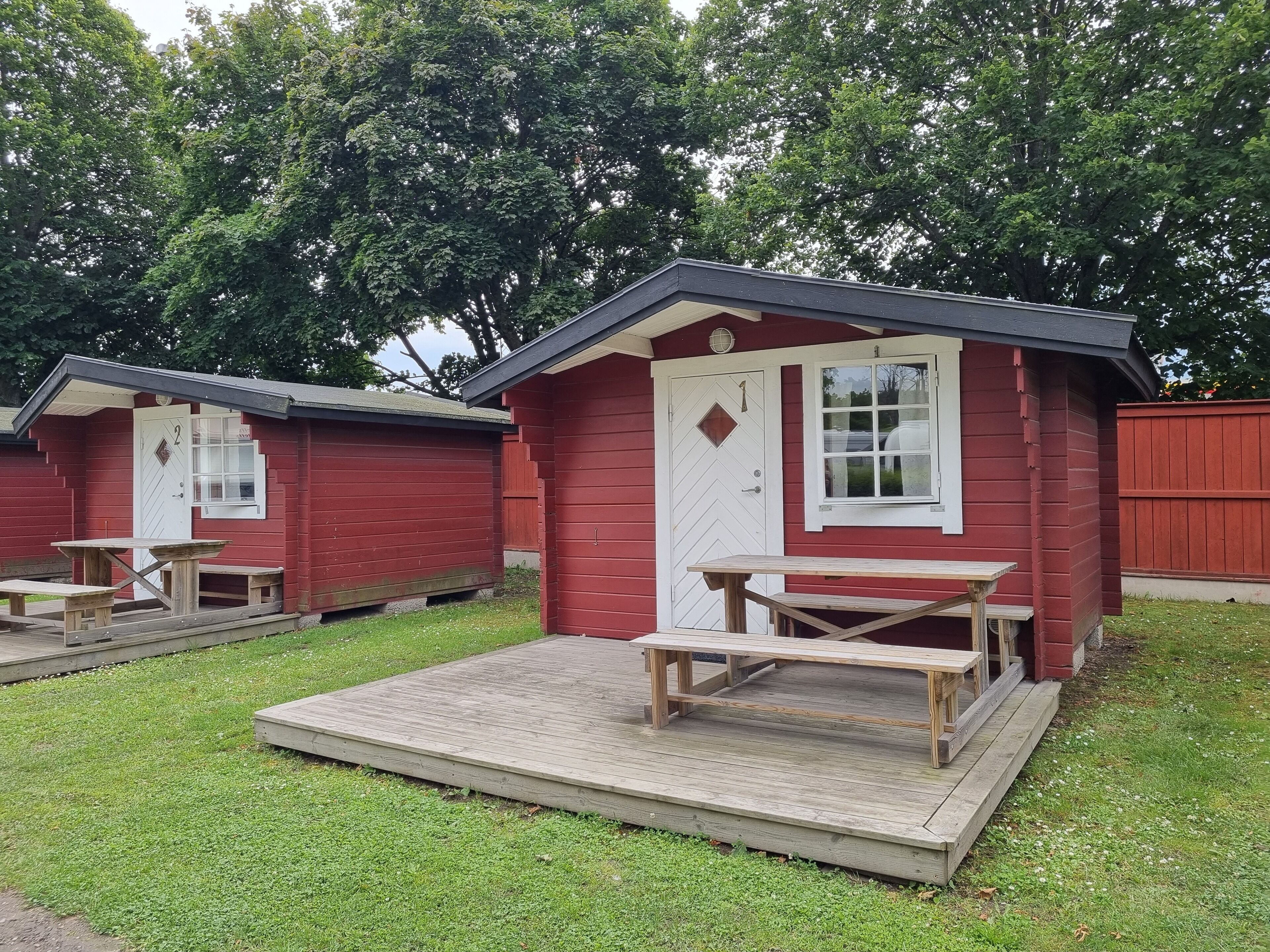 A red wooden cabin with a white door and window, a wooden bench, and a picnic table on a grassy area.