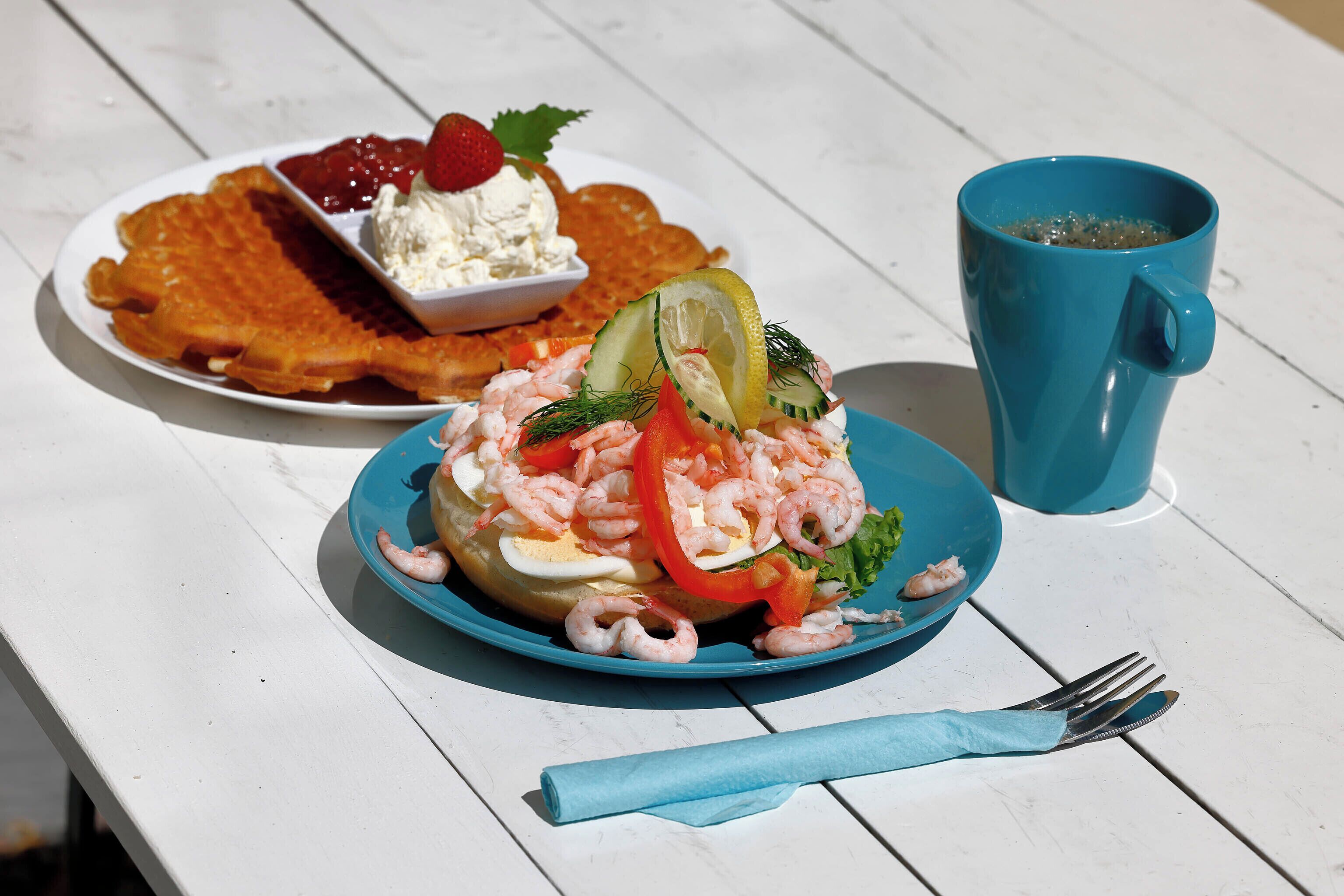 A meal with shrimp, eggs, and vegetables on a blue plate, a bowl of dessert, and a blue cup on a white table.