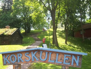 Un letrero de madera con el nombre 'Korskullen' en letras grandes, un edificio de piedra, una estructura roja y un camino que sube.
