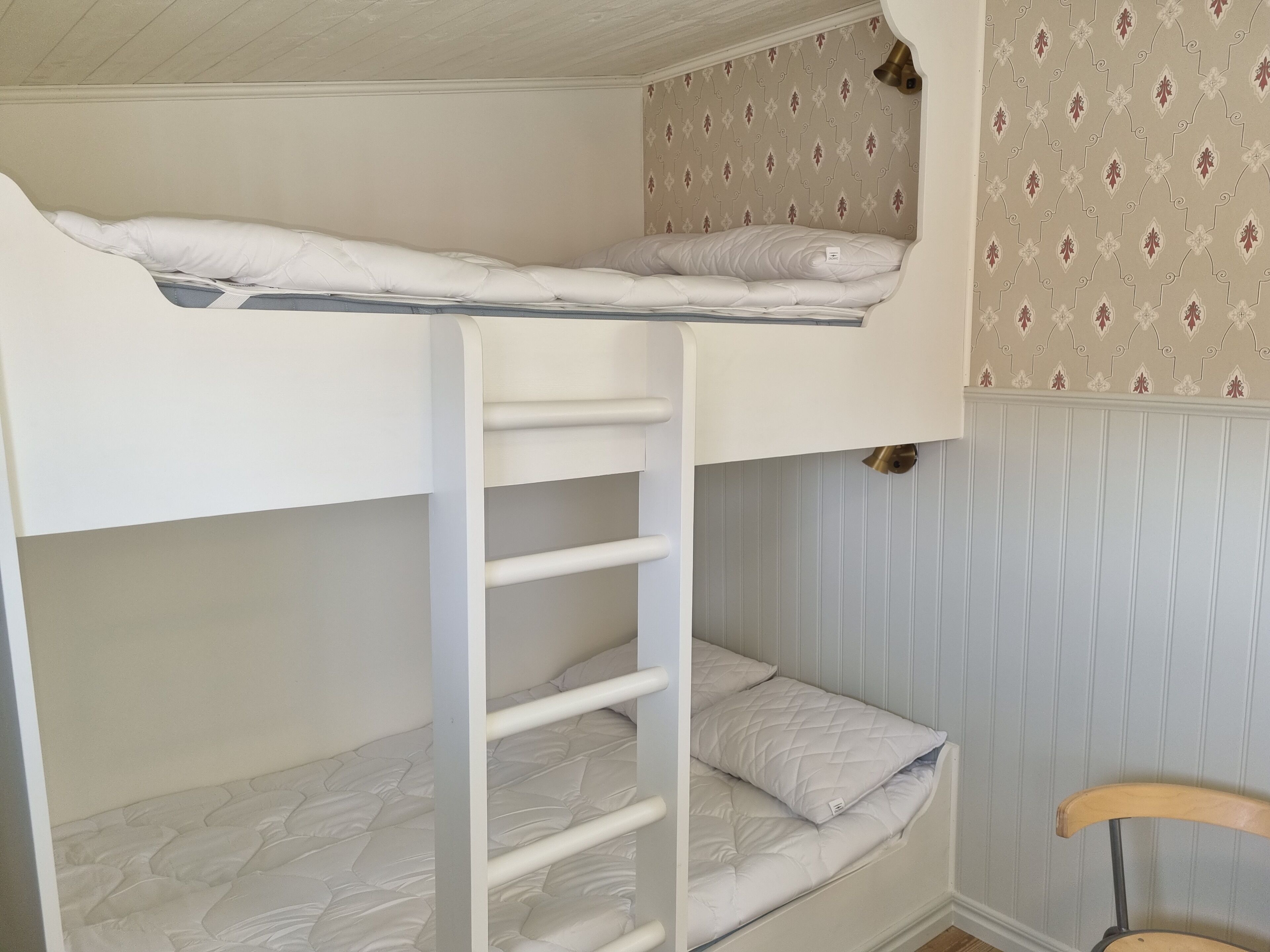 A bunk bed with a ladder, a chair, and patterned wallpaper.