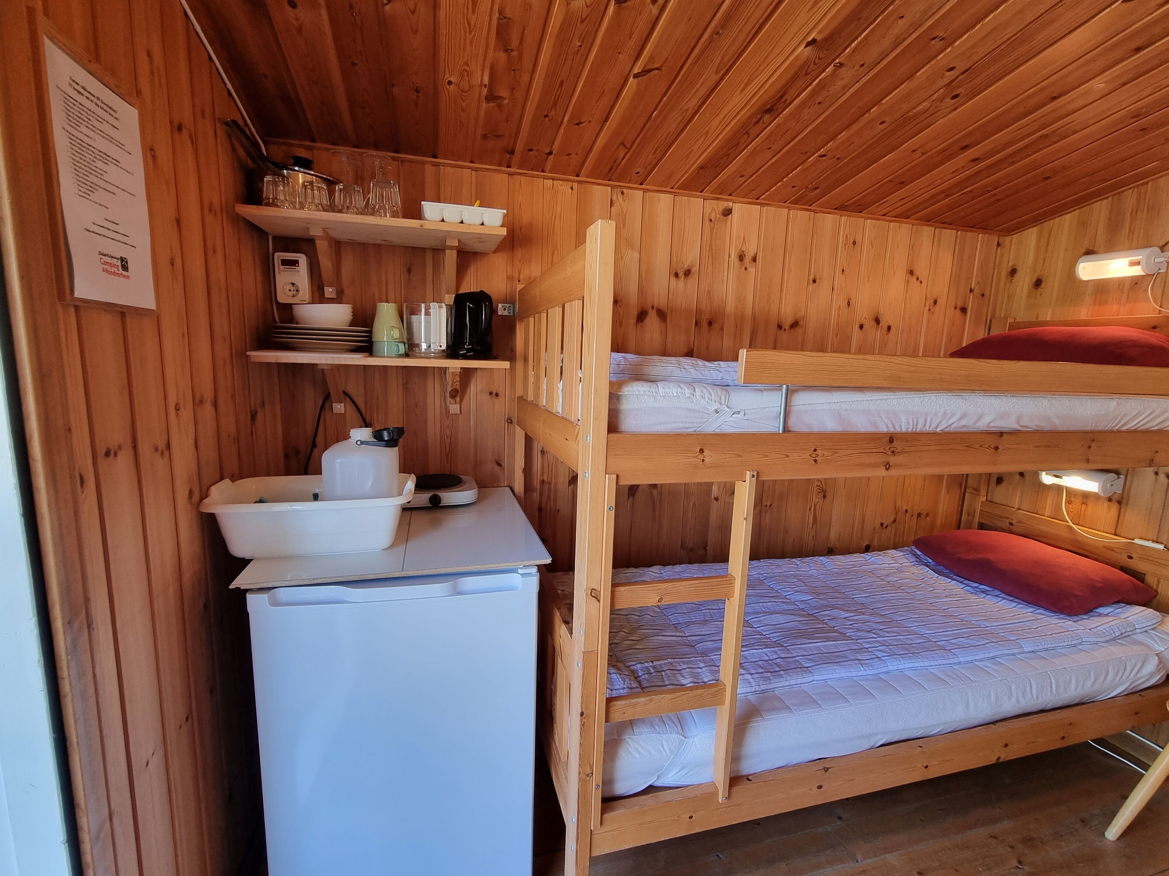 Un letto a castello con materasso bianco e cuscino rosso, un frigorifero bianco e mensole in legno con oggetti da cucina.