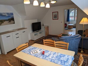 Un salon confortable avec un téléviseur, une table à manger recouverte d’une nappe à motifs et un canapé bleu.