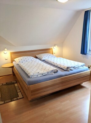 Une chambre individuelle avec un lit en bois, deux tables de chevet, une petite table d’appoint, une fenêtre avec des rideaux et un petit tapis.