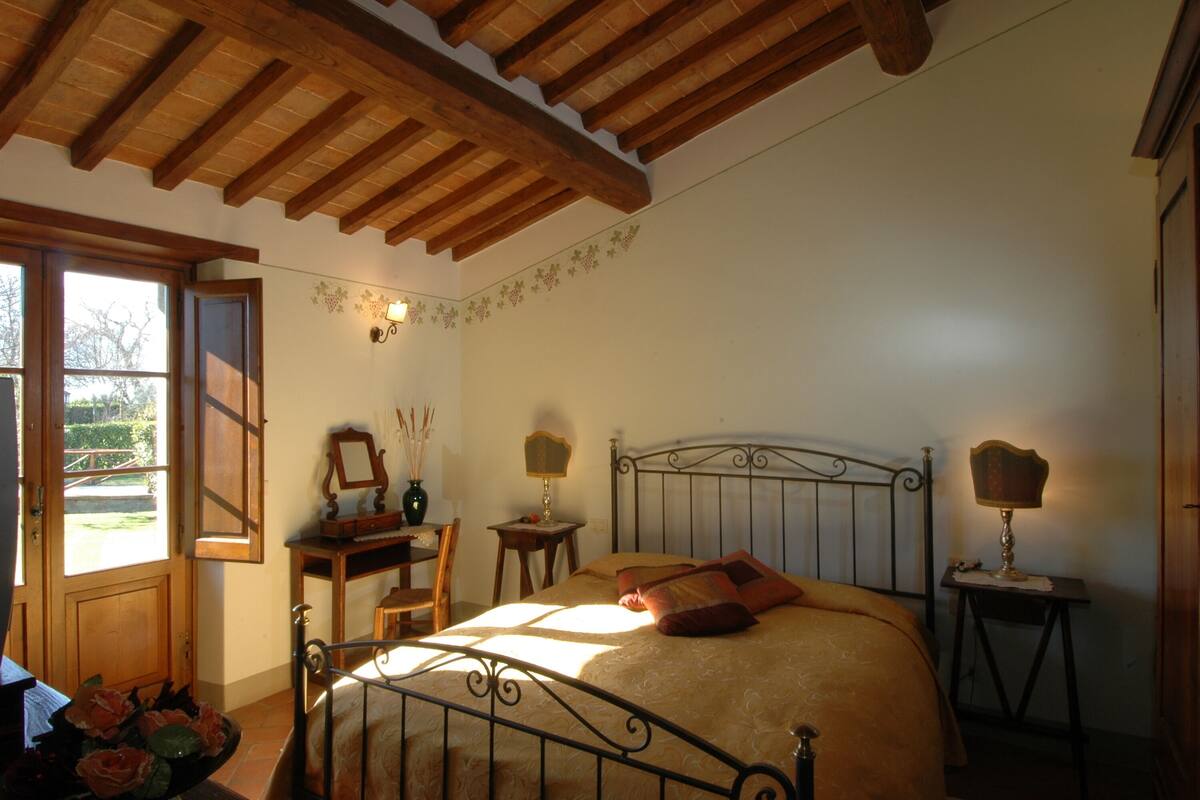 Een slaapkamer met een houten plafond, een bed met metalen frame, twee nachtkastjes met lampen, een stoel en een raam met uitzicht op groen.