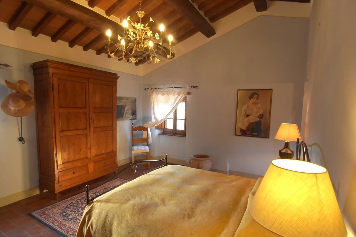 Een slaapkamer met een bed, een houten kledingkast, een kroonluchter, een stoel, een tafel en een lamp.