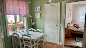 A room with a wooden floor, white walls, a white door, a dining table with chairs, a window with a checkered curtain, and a bed with a wooden headboard. - Skeppsdockans Camping & Vandrarhem (Soderkoping)