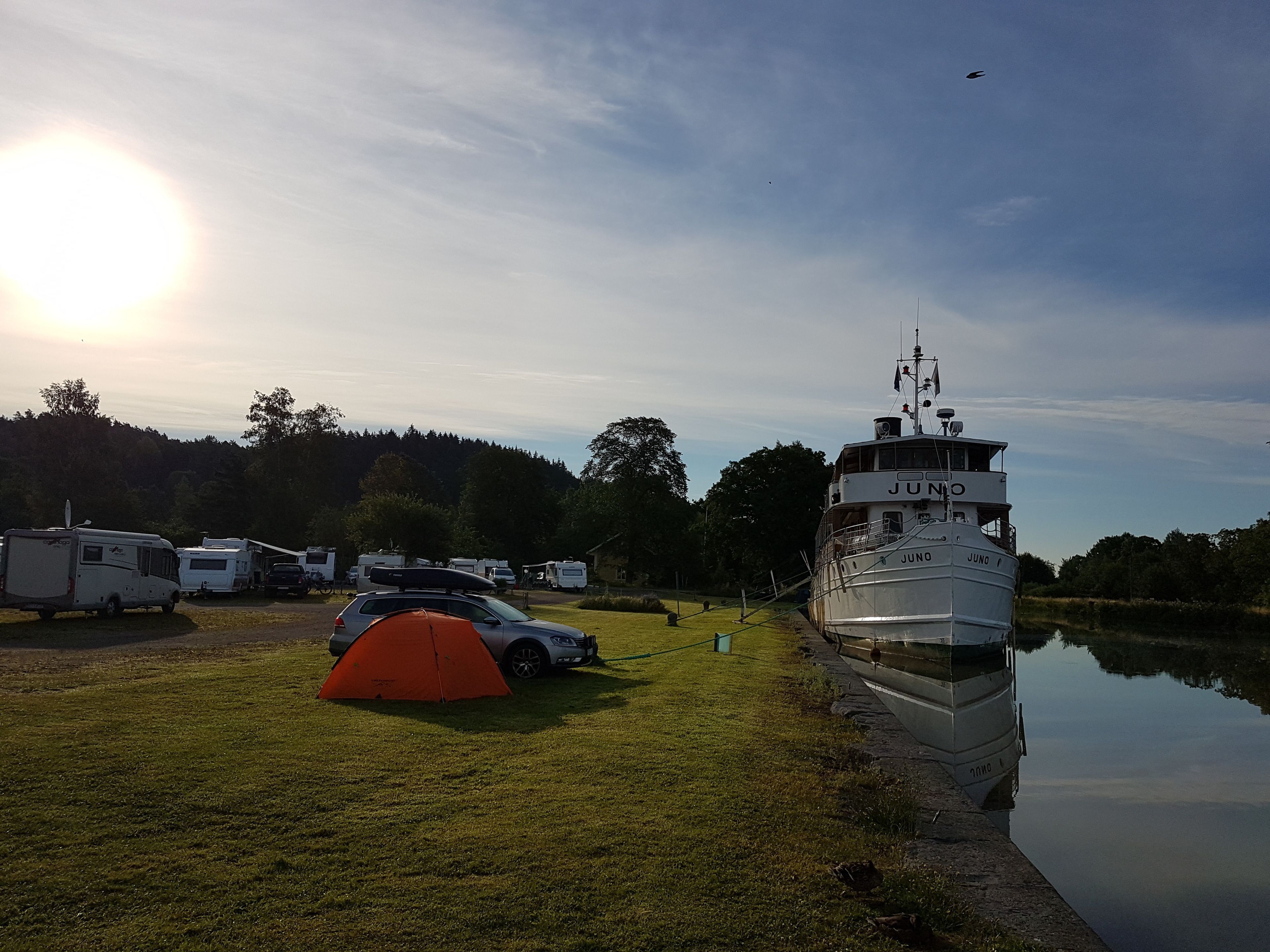 Een camping met campers en een tent, een boot genaamd Juno afgemeerd aan het water, en een heldere lucht.