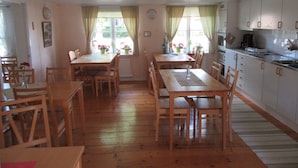 Un coin repas avec des tables et des chaises en bois, une cuisine avec des meubles blancs et une fenêtre avec des rideaux.