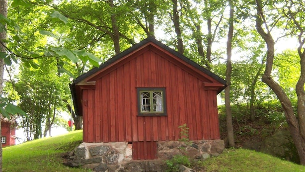 Une cabane en bois rouge, posée sur des fondations en pierre, entourée d’une végétation luxuriante et d’arbres.