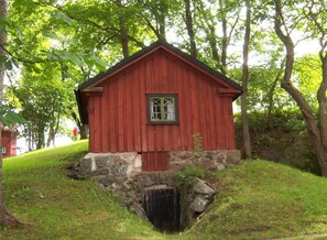 Une cabane en bois rouge, posée sur des fondations en pierre, entourée d’une végétation luxuriante et d’arbres.
