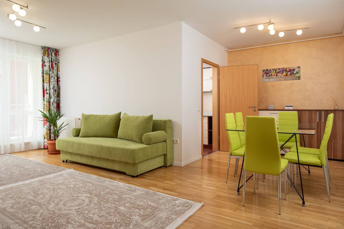 A modern living room with a green sofa, a dining area with bright green chairs, and a kitchen visible through an open door.