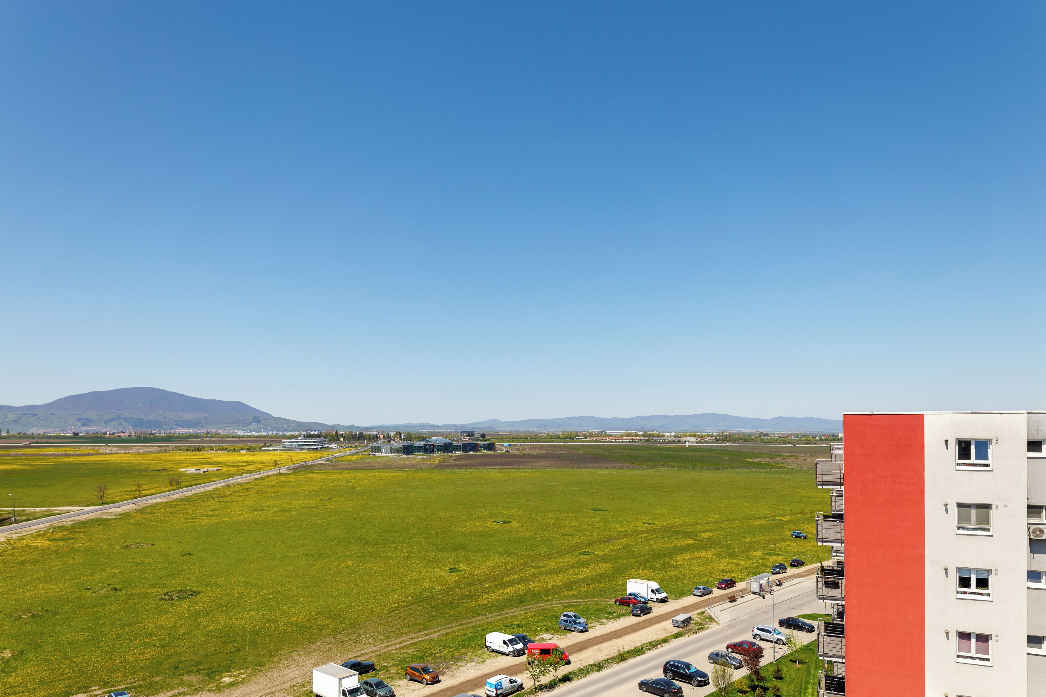 Un immeuble de grande hauteur à la façade rouge, un parking où sont stationnés divers véhicules, et un terrain verdoyant avec une montagne en arrière-plan.
