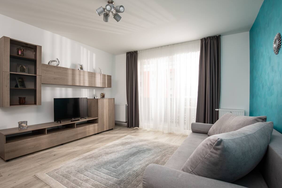 A modern living room with a flat-screen TV on a wooden TV stand, a grey sofa, and a large window with sheer curtains.