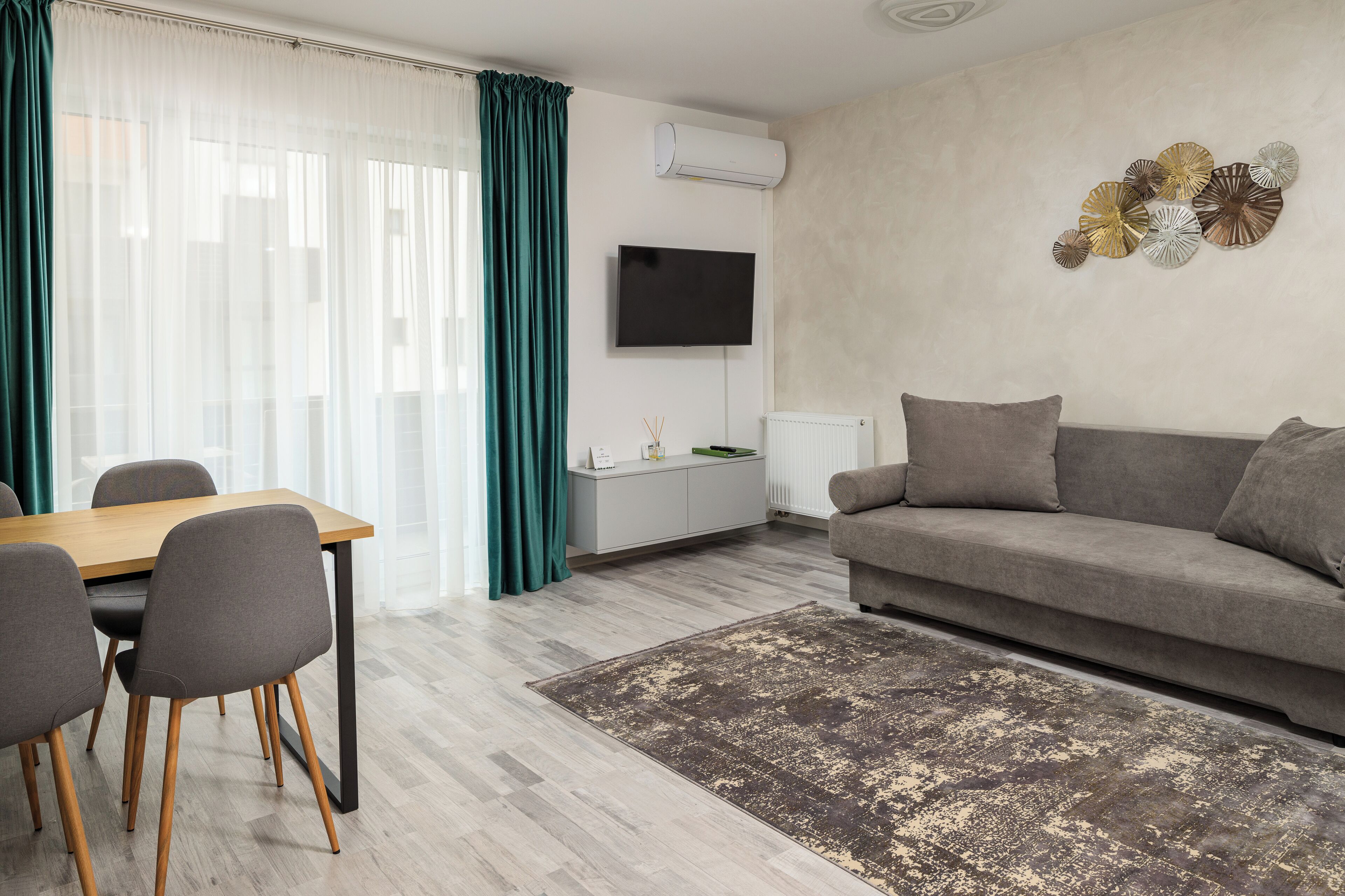 A modern living room with a wooden dining table, grey chairs, a grey sofa, a patterned rug, and a wall-mounted TV.