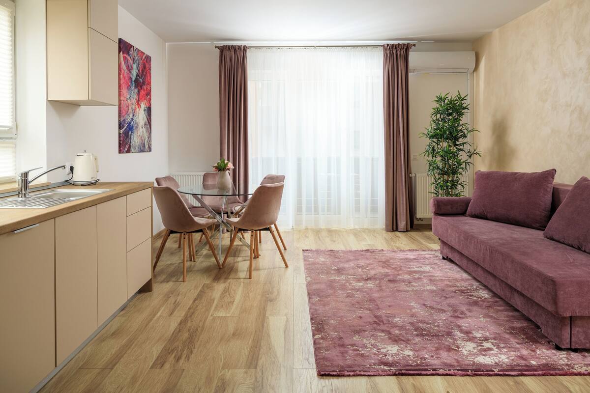 A modern living room with a wooden floor, a glass dining table with chairs, a pink sofa, and a large window with curtains.