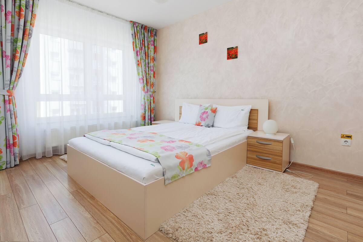 A bedroom with a bed, a bedside table, a nightstand, a fluffy rug, and a window with curtains.