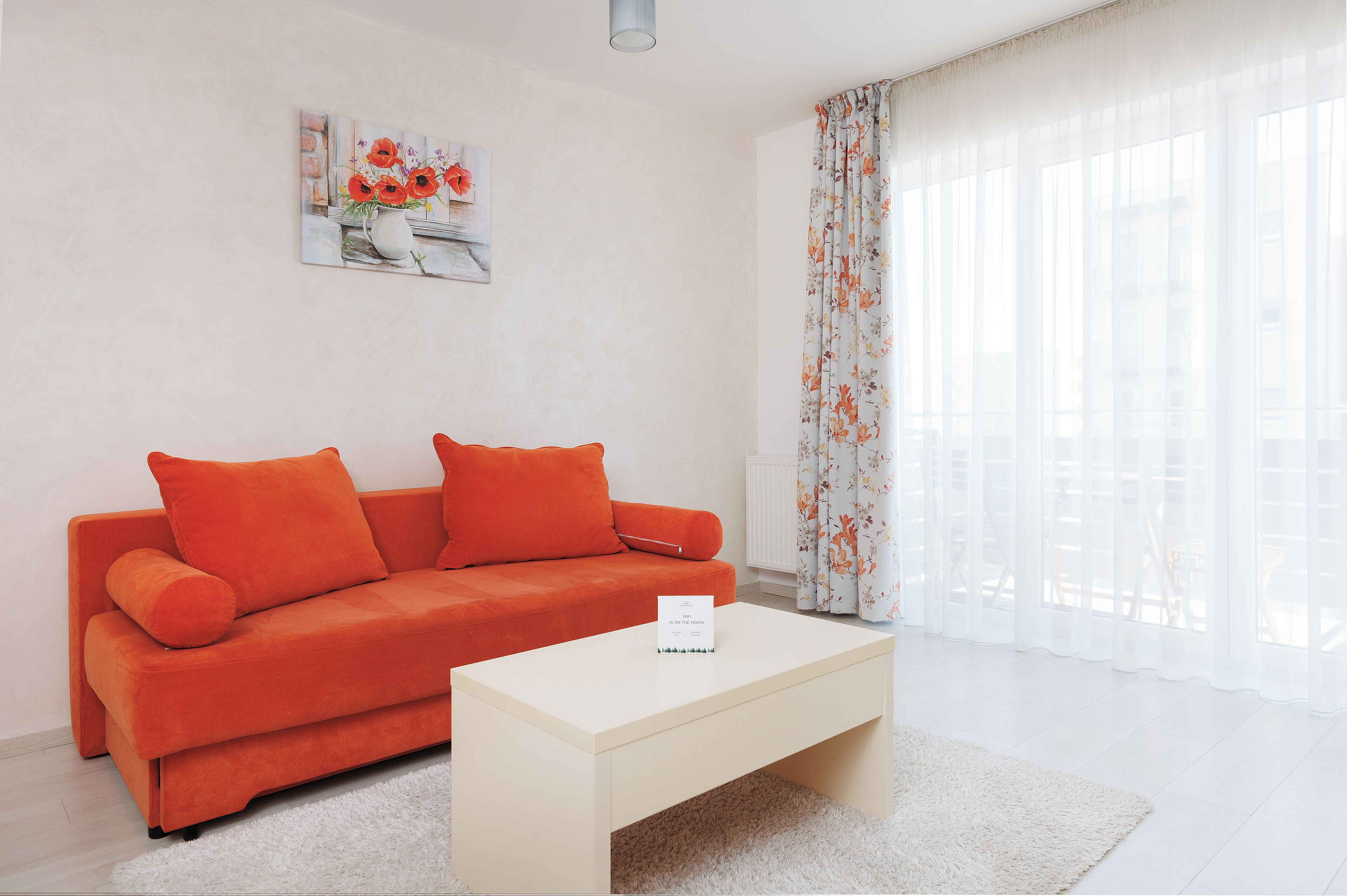 A modern living room with a bright orange sofa, a white coffee table, and a floral curtain.