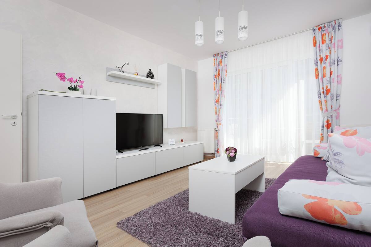 A modern living room with a flat-screen TV, white furniture, a purple sofa, and a floral curtain.