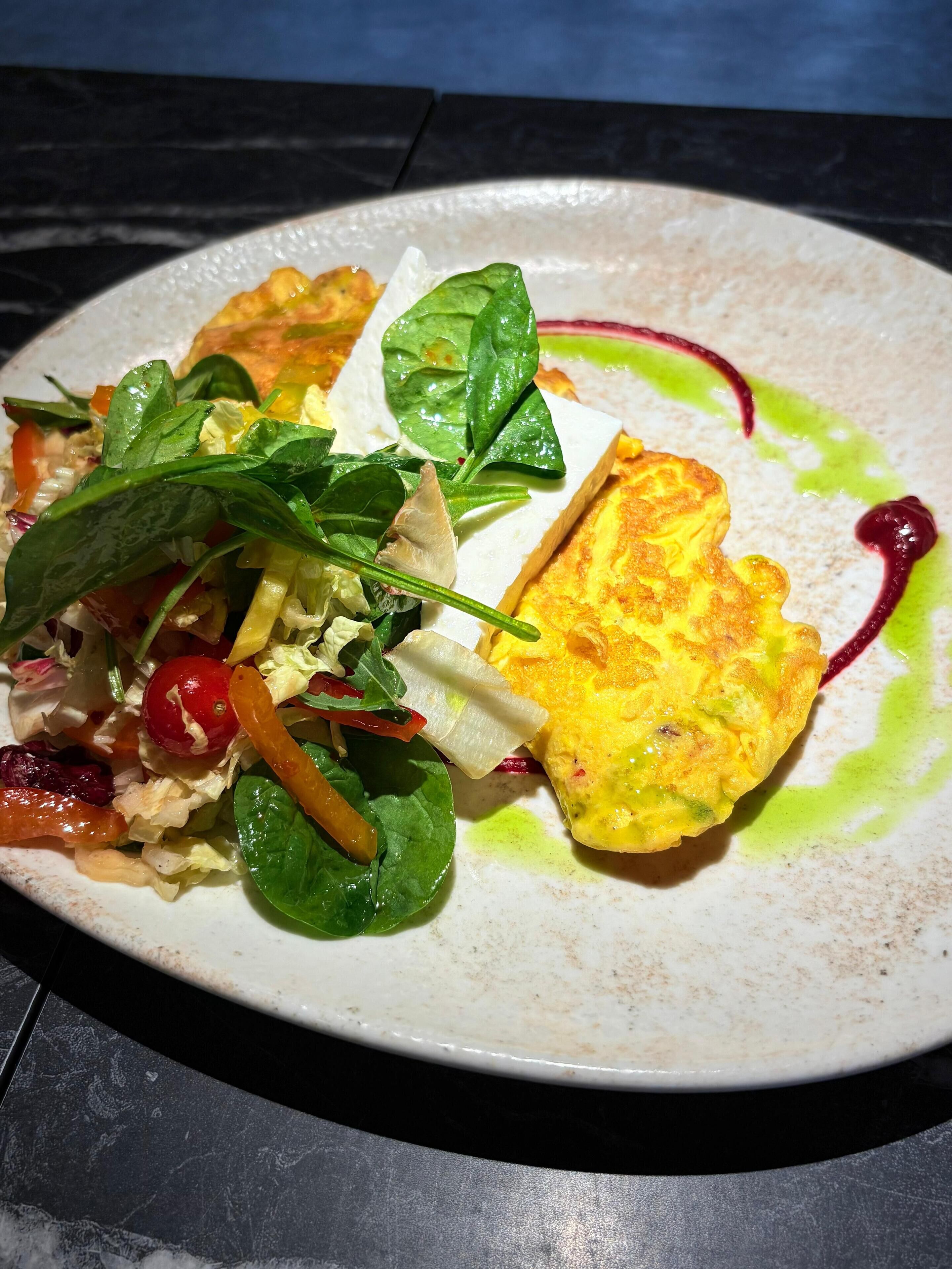 Um prato de comida com salada e um alimento frito, possivelmente uma omelete, sobre um prato branco com molho verde.