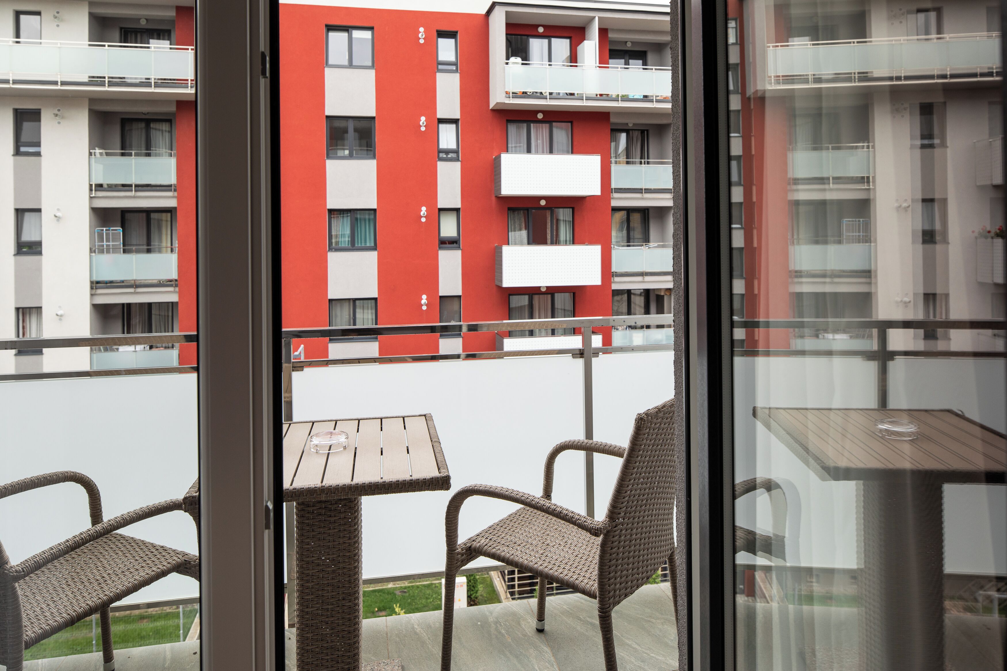 Een balkon met rieten stoelen en een tafeltje, met uitzicht op een gebouw met rode en witte buitenmuren.