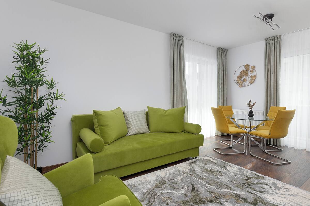 A living room with a green sofa, yellow chairs, a glass coffee table, and a large plant.