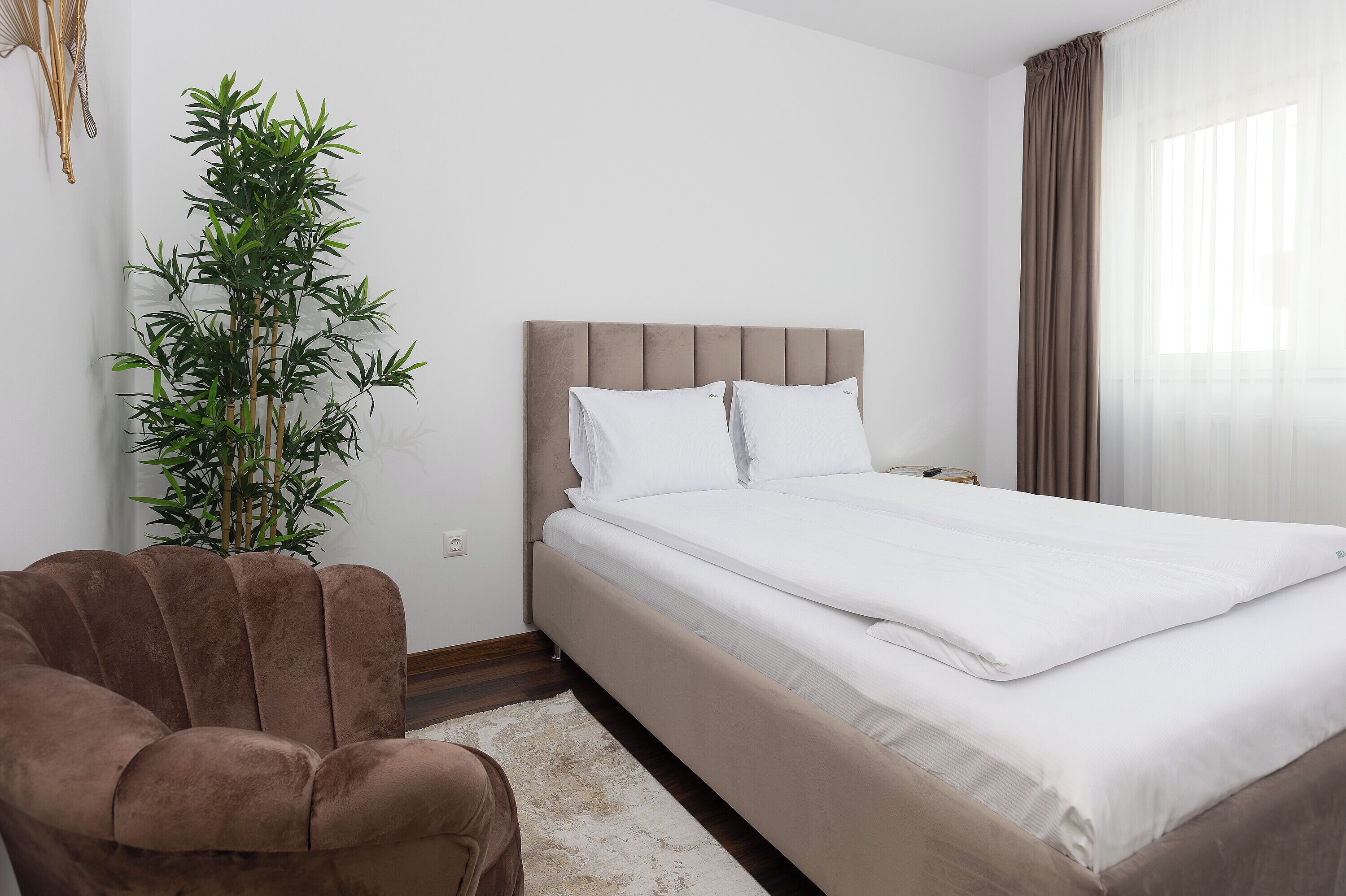 A neatly made bed with white linens, a brown upholstered chair, and a potted plant.